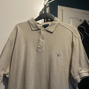 Beige pikétröja från GANT - Säljer en stilren beige pikétröja från GANT. Tröjan har en klassisk krage med knappar och det ikoniska GANT-emblemet broderat på bröstet. Perfekt för sommarn 