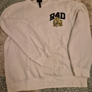 Vit sweatshirt med tryck - Säljer en vit sweatshirt med stort svart och gult tryck av texten 'R4D' och en ros på både fram- och baksidan. Tröjan har långa ärmar och en rund halsringning. Perfekt för en avslappnad stil.