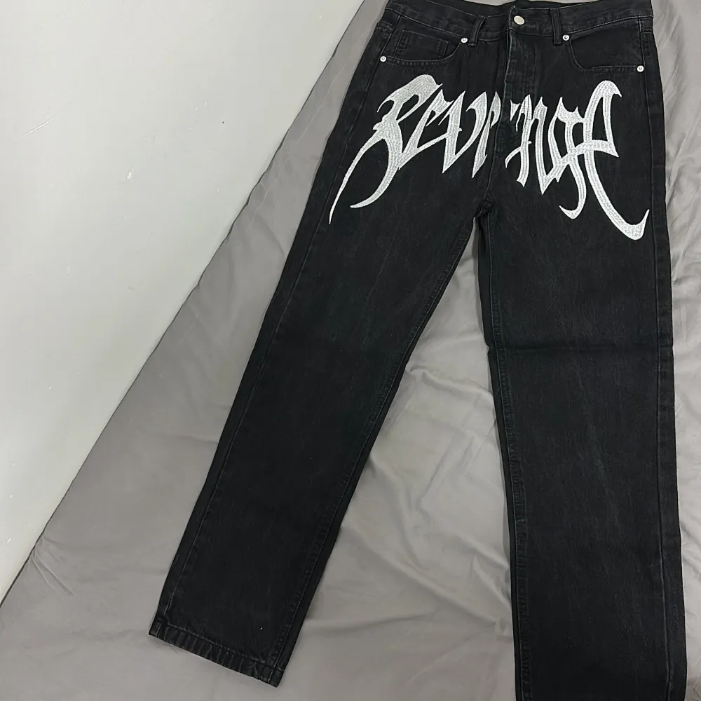 Coola svarta jeans med ett stort vitt tryck på framsidan. De har en klassisk femficksdesign och knappgylf. Perfekta för en unik look. Pris kan diskuteras.. Farkut & Housut.
