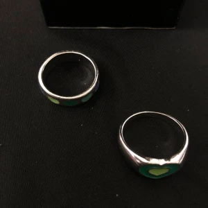 Silverringar med hjärtmönster - Två stilrena silverringar med färgglada hjärtan. Den ena ringen har ett stort grönt hjärta i mitten, medan den andra har flera små hjärtan i grönt och blått runt om. Perfekta för att ge en färgklick till din stil.