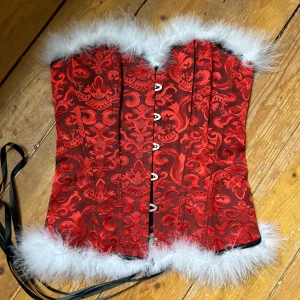 Röd korsett med pälsdetaljer - Snygg röd korsett med ett elegant mönster och vita pälsdetaljer. Den har snörning bak och knäppning fram, vilket ger en perfekt passform. Perfekt för att sticka ut vid speciella tillfällen.