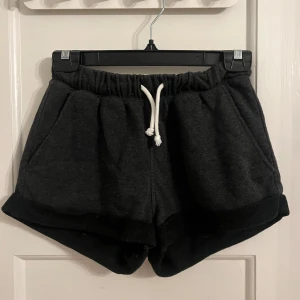 Mörkgrå mjukisshorts med snörning - Säljer ett par mörkgrå mjukisshorts med elastisk midja och vit snörning. Shortsen har en avslappnad passform och är perfekta för en bekväm stil. De har också praktiska fickor på sidorna.