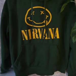 Grön Nirvana hoodie från JHK - Säljer en grön hoodie med Nirvana-tryck i gult från JHK. Hoodien har en klassisk passform med känguruficka och justerbar huva. Perfekt för fans av bandet! Tillverkad i en blandning av polyester och bomull.
