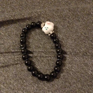Svart pärlarmband med en get - Charmigt svart pärlarmband med en unik vit figur i mitten. Figuren har små färgdetaljer i grönt och rött, vilket ger en lekfull touch. Perfekt för att lägga till en personlig stil till din outfit.