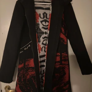 Svart kappa från Desigual - Snygg svart kappa från Desigual med unikt rött och svart mönster på insidan. Jackan har långa ärmar och en stilren design som passar perfekt för höst och vinter. Den har en dragkedja framtill och är tillverkad i ett mjukt material för extra komfort.