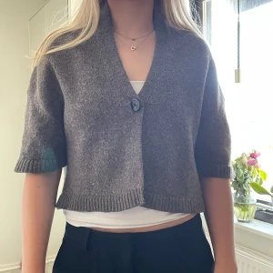 Grå kofta med korta ärmar - Säljer en stilren grå/brun kofta med korta ärmar och en knapp framtill. Koftan är croppad och har ribbade kanter som ger en snygg detalj. Perfekt för lager-på-lager-stil.