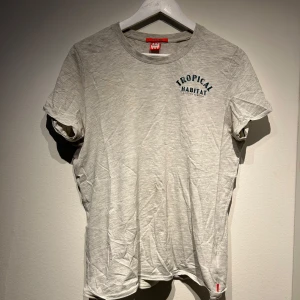 Grå t-shirt med tryck 'Tropical Habitat' - Säljer en grå t-shirt med texten 'Tropical Habitat' tryckt på bröstet och ett stort färgglatt snäckmotiv på baksidan. T-shirten har en rund hals och korta ärmar. Perfekt för en avslappnad stil.