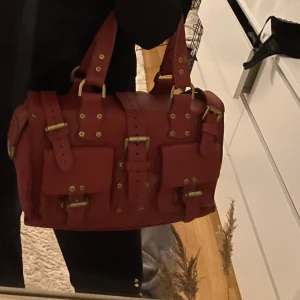 Röd skinnväska från Mulberry - Säljer en snygg röd skinnväska från Mulberry med flera spännen och detaljer i metall. Väskan har två robusta handtag och en praktisk dragkedja för kort. Perfekt för dig som vill ha en stilren och rymlig väska med karaktär. Tar gärna emot prisförslag 