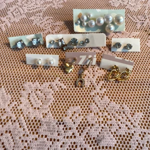 Begagnade örhängen  - Ett set med flera olika örhängen, inklusive pärlor, guldiga hjärtan och hästskor, samt glittriga stenar i olika former. De som sitter flera på rad är parlösa.🌸