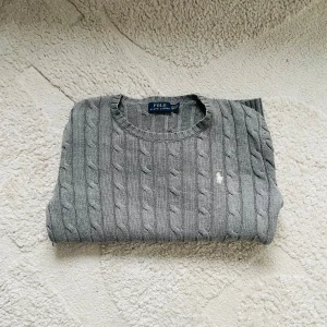 Grå stickad tröja från Ralph Lauren - Säljer en stilren grå stickad tröja från Ralph Lauren med kabelstickat mönster. Tröjan har en rund halsringning och långa ärmar. 