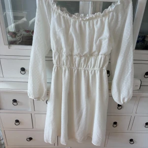 Vit  klänning - Charmig vit offshoulder klänning med elastisk midja och långa ärmar. Klänningen har ett diskret prickigt mönster och volangdetaljer vid halsringningen. Perfekt för sommarens alla tillfällen. 