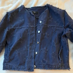 Mörkblå jacka med stjärnmönster - Säljer en unik mörkblå jacka med stjärnmönster och fransiga kanter. Jackan har knappar framtill och en cool, avslappnad stil. Perfekt för att ge din outfit en extra touch ⭐️ Den är liten i strl så skulle säga den passa mer S än M 