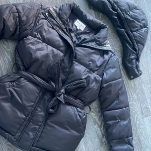 Svart pufferjacka från NA-KD - Säljer en svart pufferjacka från NA-KD med avtagbar huva och knytbälte i midjan. Jackan har dragkedja och tryckknappar framtill för extra skydd mot kyla. Perfekt för kalla dagar! 🖤Oversized i storleken och det finns några sminkfläckar på insidan av kragen men skulle inte säga att det syns mycket alls. 