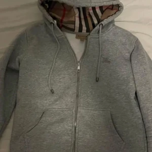 Grå huvtröja med dragkedja - Säljer en stilren grå huvtröja från Burberry med dragkedja och klassiskt rutigt mönster i huvan. Tröjan har långa ärmar och praktiska fickor framtill. Perfekt för en avslappnad look.