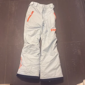 Ljusblå täckbyxor från Helly Hansen - Snygga ljusblåa täckbyxor från Helly Hansen med orangea dragkedjor och detaljer. Perfekta för vinterns äventyr med en bekväm passform och slitstarkt material. Byxorna har justerbar midja och praktiska fickor med dragkedja. 