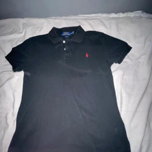 Svart pikétröja från Ralph Lauren - Säljer en klassisk svart pikétröja från Ralph Lauren i slim fit. Tröjan har en liten röd logga på bröstet och knappar framtill. Perfekt för en stilren look.