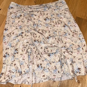 Blommig kjol med volangdetaljer - Charmig blommig kjol i beige med blå och bruna blommor. Kjolen har volangdetaljer och snörning framtill som ger en romantisk touch. Perfekt för en somrig look! I storlek S