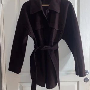 Mörk lila kappa från Gina Tricot - Snygg brun kappa från Gina Tricot i storlek XL. Den har en klassisk krage, knappar framtill och ett bälte i midjan för en stilren look. Perfekt för höst och vår. Köpt förra våren för 599kr