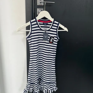 Randig ärmlös klänning från GANT - Snygg ärmlös klänning från GANT med marinblå och vita ränder. Klänningen har en volangdetalj vid fållen och ett stilrent 'G' emblem på bröstet. Perfekt för en avslappnad och stilren look.