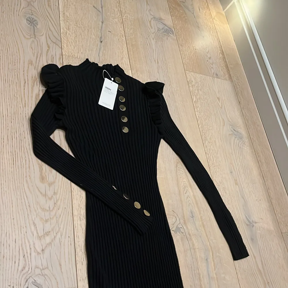 Snygg svart ribbad klänning från Moocci med långa ärmar och dekorativa guldknappar framtill. Perfekt för en stilren look med en touch av elegans. Klänningen har en hög krage och volangdetaljer vid axlarna. Ett måste i garderoben för den modemedvetna! 🖤. Mekot.