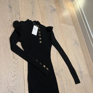 Svart ribbad klänning från Moocci - Snygg svart ribbad klänning från Moocci med långa ärmar och dekorativa guldknappar framtill. Perfekt för en stilren look med en touch av elegans. Klänningen har en hög krage och volangdetaljer vid axlarna. Ett måste i garderoben för den modemedvetna! 🖤