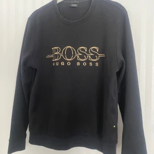 Svart tröja från Hugo Boss - Snygg svart tröja från Hugo Boss med guldig logga på bröstet. Tröjan har en slim fit passform och långa ärmar. Perfekt för en stilren look. Den är som helt ny men säljs för att den inte passar min stil längre. Pris går alltid att diskuteras.