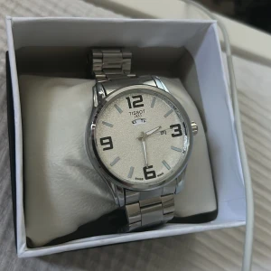 Silverfärgad klocka från Tissot - Elegant silverfärgad klocka från Tissot med tydlig urtavla och stora siffror. Klockan har ett robust armband i metall och klassisk design. Perfekt för den stilmedvetna som vill ha en tidlös accessoar.