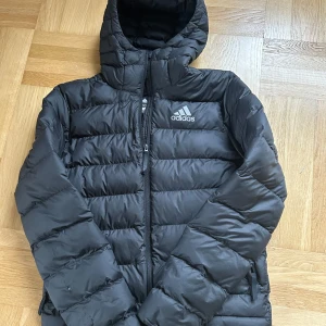 Svart pufferjacka från Adidas - Säljer en svart pufferjacka från Adidas med dragkedja och huva. Jackan har en broderad logga på bröstet och är perfekt för kyligare väder. Den är långärmad och har en stilren design. Liten defekt på bild 3.