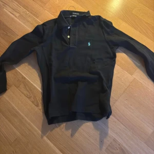 Svart pikétröja från Ralph Lauren - Snygg svart pikétröja från Ralph Lauren med klassisk krage och knappar. Den har en liten grön logga på bröstet och långa ärmar. Perfekt för en stilren look. BARN STORLEK:S
