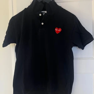 Mörklblå pikétröja med hjärtdetalj - Snygg blå pikétröja från Comme des Garçons Play med ett ikoniskt rött hjärta på bröstet. Tröjan har korta ärmar och knappar framtill. Perfekt för en stilren look.