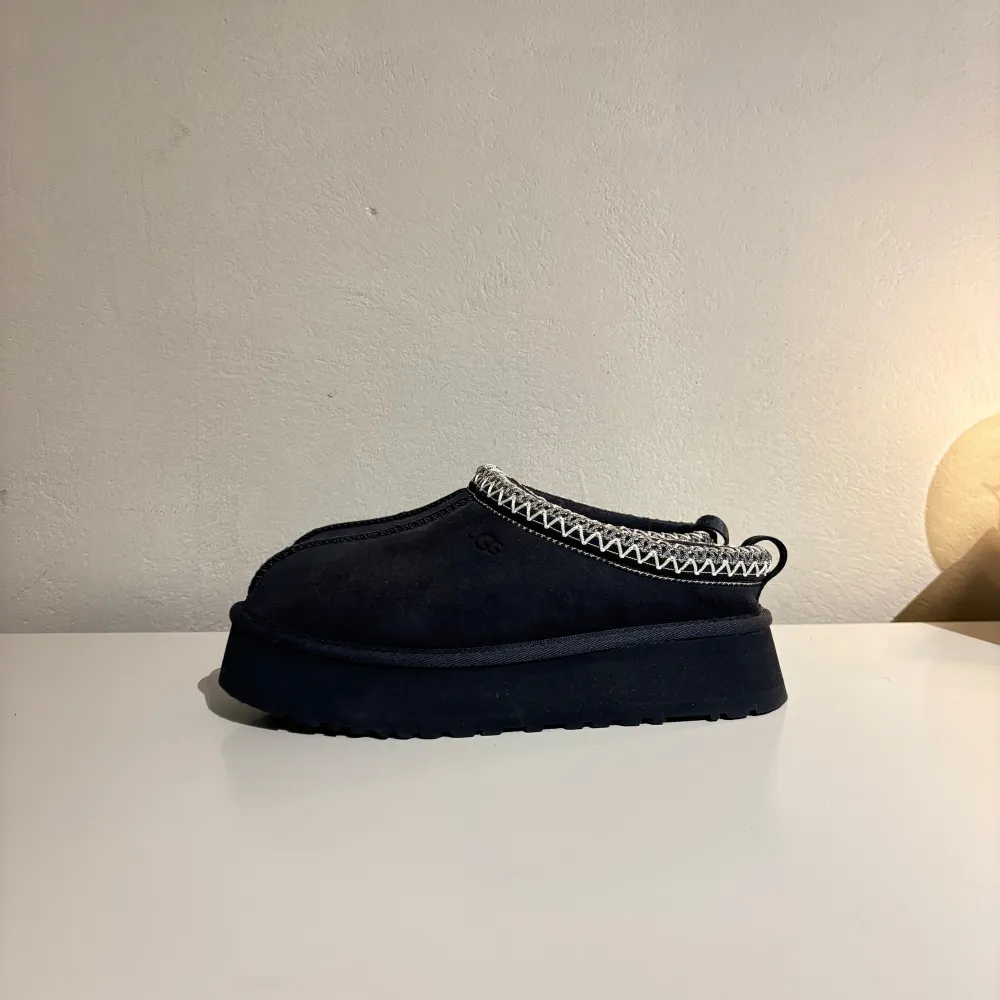 Säljer ett par mörkblå mockaskor från UGG med dekorativ kantdetalj. Skorna har en robust sula och är perfekta för kyligare dagar. De är bekväma och stilrena, idealiska för både inomhus och utomhusbruk.. Kengät.