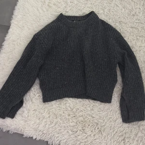 Mörkgrå stickad tröja - Säljer en mysig mörkgrå stickad tröja med ribbade detaljer. Tröjan har en croppad passform och långa ärmar, perfekt för kyliga dagar. Den är både stilren och bekväm, ett måste i garderoben! 🖤
