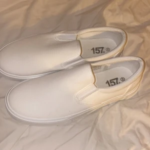 Vita slip-on sneakers från 157 - Snygga och enkla vita slip-on sneakers från 157. Perfekta för en avslappnad stil med en bekväm passform. Skorna har en klassisk design med vit ovandel och bruna gummisulor. OBS! Oanvända