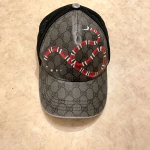Grå keps med ormmönster från Gucci - Snygg grå keps från Gucci med ett iögonfallande ormmönster i rött och vitt. Kepsen har ett klassiskt Gucci-mönster och justerbar rem baktill. Perfekt för att ge din outfit en lyxig touch.