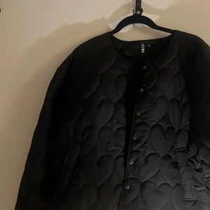 Svart quiltad jacka från Pieces - Säljer en svart quiltad jacka från Pieces med hjärtmönster. Jackan har knappar framtill och en rund halsringning. Perfekt nu till våren