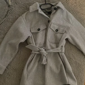 Grå kappa från Zara - Säljer en snygg grå kappa från Zara i storlek S. Den har långa ärmar och ett bälte i midjan för en fin passform. Perfekt för höst och vår! Kappan har knappar framtill och två bröstfickor med lock. Superfin och stilren design!