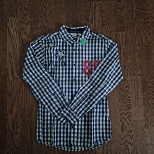 Rutig skjorta för Pojkar - Säljer en cool rutig skjorta från Lindex i storlek EUR 164. Skjortan är svart och vit med långa ärmar och knappar. Den har snygga detaljer som en stjärna och siffran 89 i rött framtill, samt stort tryck på ryggen. Perfekt för höst och vår. Säljer för 99:-