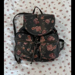 Blommig Ryggsäck - Blommig ryggsäck i vinyl. Två små fack på utsidan och två på insidan. Knappen som stänger stora facket sitter   löst, går att sätta fast om man vill (syns på bild 3), i övrigt är väskan i fint skick. Stora facket stängs med snörning samt knapp.