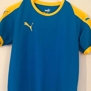 Blå och gul fotbollströja från Puma - Säljer en snygg blå fotbollströja från Puma med gula detaljer på ärmarna och axlarna. Tröjan är kortärmad och perfekt för både träning och match. Den är i bra skick och redo för nya äventyr på fotbollsplanen. Passar perfekt för dig som gillar en sportig stil! ⚽️