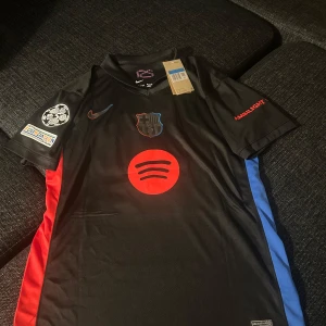 Barcelona borta tröja med lamine yamal - Snygg svart fotbollströja från Nike med korta ärmar. Tröjan har ett rött och blått klubbmärke på bröstet och röda detaljer. På ärmen finns UEFA Foundation-märke. Perfekt för fotbollsfans! Lamine yamal tröja i storlek M och det är en replika helt ny med tagg!!!