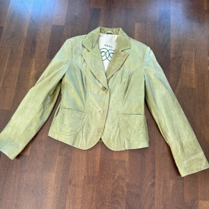 Grön mockajacka från Saki - Snygg grön mockajacka från Saki med klassisk krage och två knappar framtill. Jackan har två fickor med lock och en figursydd passform som ger en elegant look. Perfekt för att lyfta din stil! Okej skick 