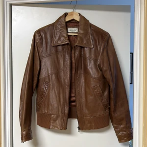 Brun skinnjacka från Hollis - Snygg brun skinnjacka från Hollis med dragkedja och ribbade detaljer vid ärmslut och nederkant. Jackan har en klassisk krage och två fickor framtill. Perfekt för en stilren look.