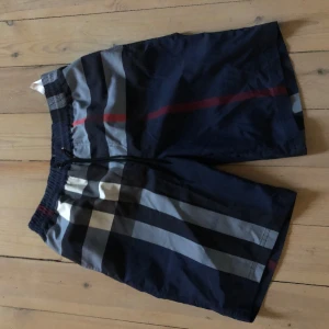 Rutig badshorts från Burberry - Snygga badshorts från Burberry i ett klassiskt rutigt mönster med nyanser av blått, grått och rött. Shortsen har en elastisk midja för bekväm passform och är perfekta för sommarens badäventyr.