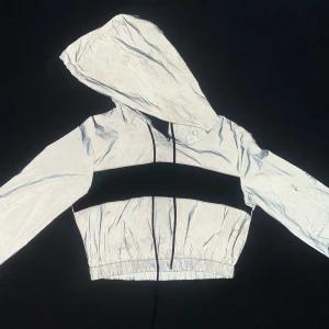 Reflekterande croppad hoodie - Snygg och modern croppad hoodie med reflekterande material och luva. Tröjan har en grå bas med en svart rand över bröstet och justerbara snören i luvan. Perfekt för att sticka ut i mängden!