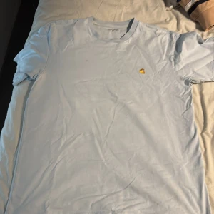 Ljusblå t-shirt från Carhartt - Säljer en stilren ljusblå t-shirt från Carhartt med en liten broderad logga på bröstet. Perfekt för en avslappnad look. T-shirten har en klassisk passform och är tillverkad i mjukt bomullsmaterial.