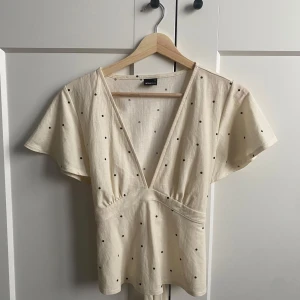 Topp med knytning från gina - Säljer en söt beige blus från gina tricot med svarta prickar. Blusen har en v-ringad design och korta ärmar. Den har även ett knytband i ryggen för en fin passform. Perfekt för en avslappnad stil. Den är även slutsåld överallt.