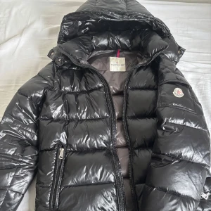 Svart pufferjacka från Moncler - Snygg svart pufferjacka från Moncler med glansig finish. Jackan har en praktisk huva och dragkedja framtill. Den är quiltad för extra värme och har Monclers logotyp på ärmen. Perfekt för kyliga dagar.