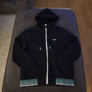 Svart hoodie från Boss - Snygg svart hoodie från Boss med dragkedja och justerbar huva. Den har långa ärmar och ett stilrent tryck med 'BOSS' längs nederkanten. Perfekt för en avslappnad stil.