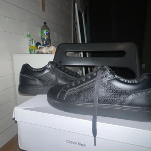 Svarta sneakers från Calvin Klein - Snygga svarta sneakers från Calvin Klein med monogrammönster på sidorna. Skorna har snörning och är tillverkade i syntetmaterial. Dragkedja på sidan för design och blir även väldigt enkelt och sätta på sig skorna. Perfekta för en stilren look. Använda endast 2 gånger