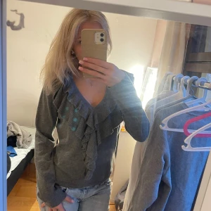 Grå tröja med volangdetaljer - Säljer en stilren grå tröja med volangdetaljer längs halsringningen. Tröjan har långa ärmar och en v-ringad design som ger en elegant touch. Perfekt för att piffa upp vardagslooken.